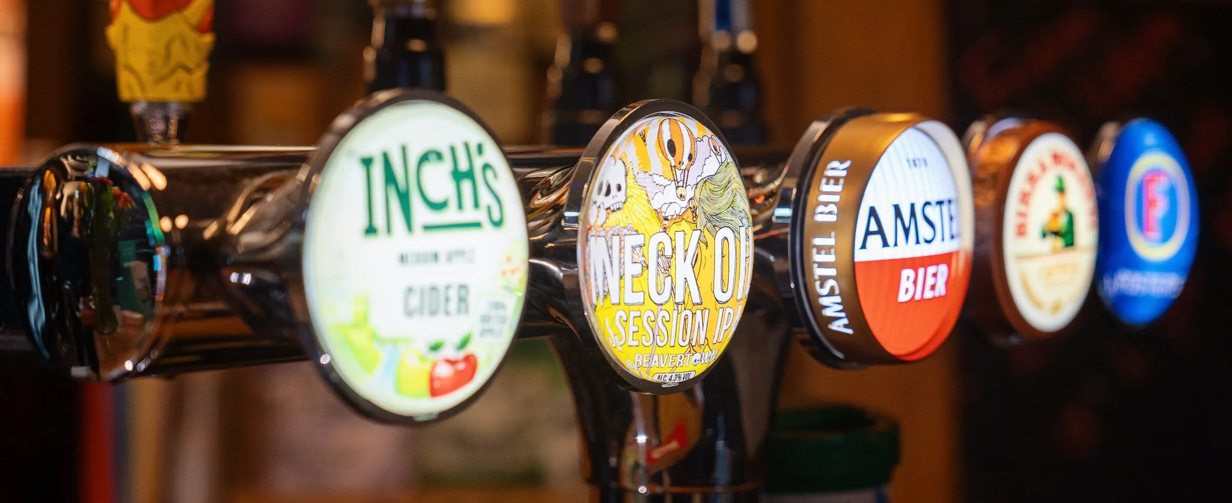 A row of beer and cider taps along the bar, their badges lit, with the back bar softly out of focus behind