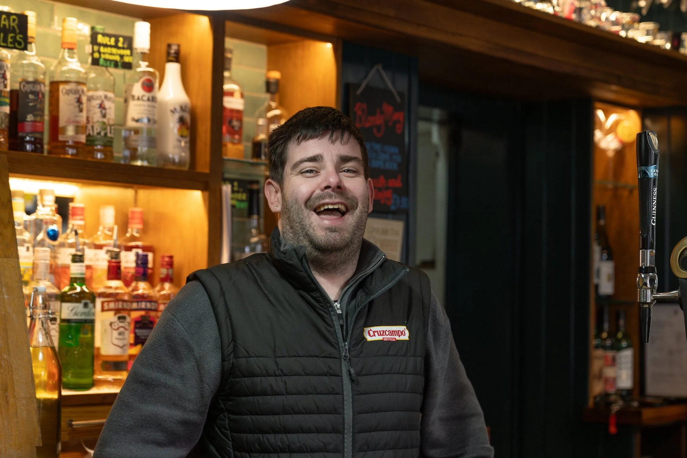 A member of the bar team laughing behind the bar, a mug of tea on the counter and the spirits shelf lit behind him