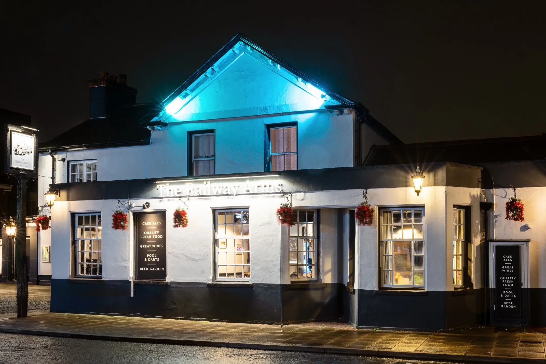 The Railway Arms at night, seen from the corner of the High Street — white frontage with dark trim, lit sign and hanging baskets
