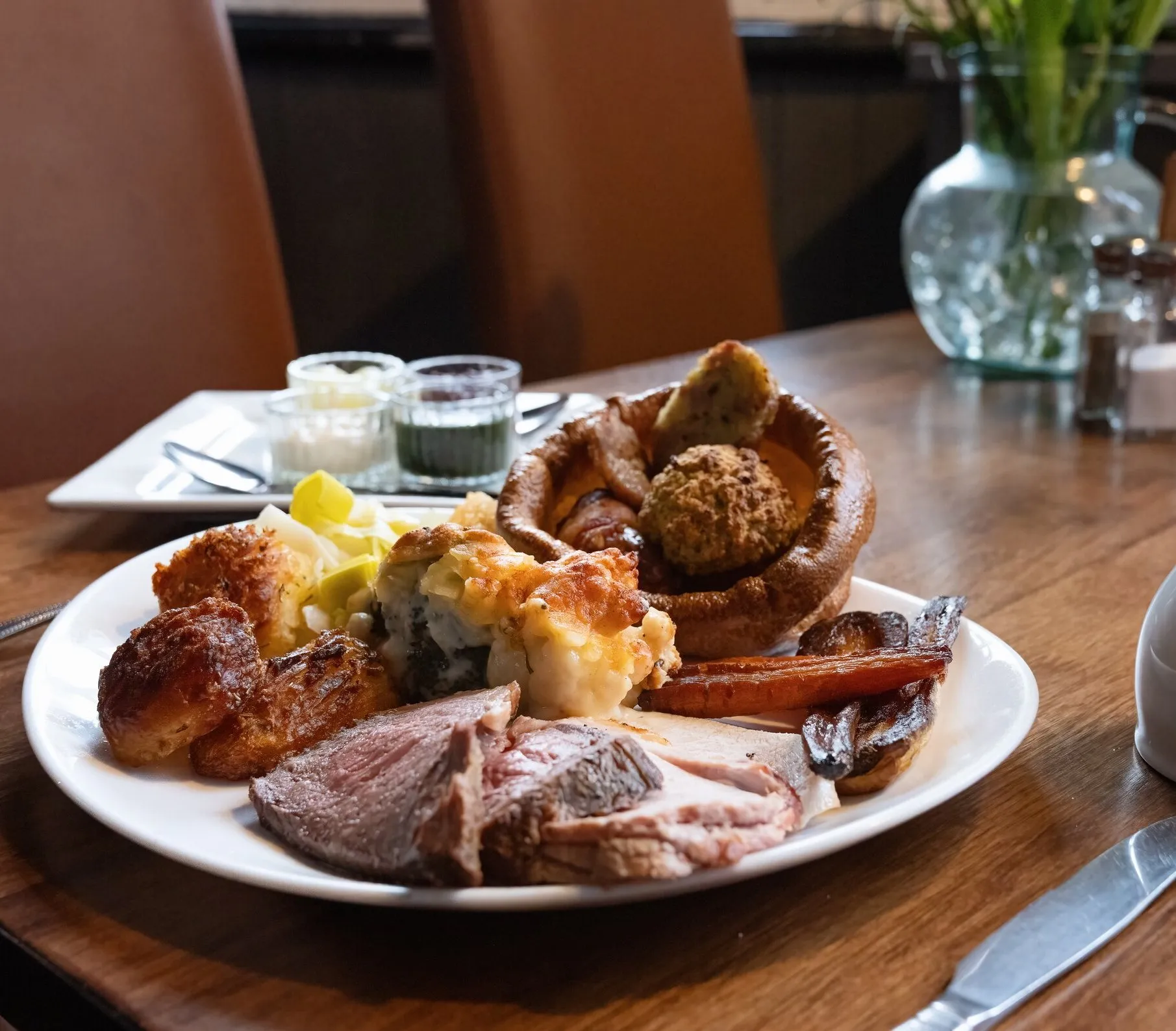 A Sunday roast on a wooden table by the window — beef, Yorkshire pudding, roast potatoes and vegetables, with a jug of gravy and a glass of wine
